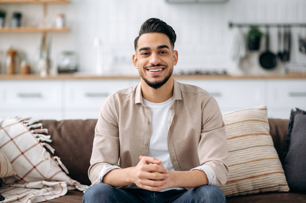 Портрет привлекательного красивого индийского или арабского парня в стильной повседневной одежде, сидящего дома на диване в гостиной, в стильном интерьере, смотрящего в камеру, положительно улыбающегося - Фото, изображение