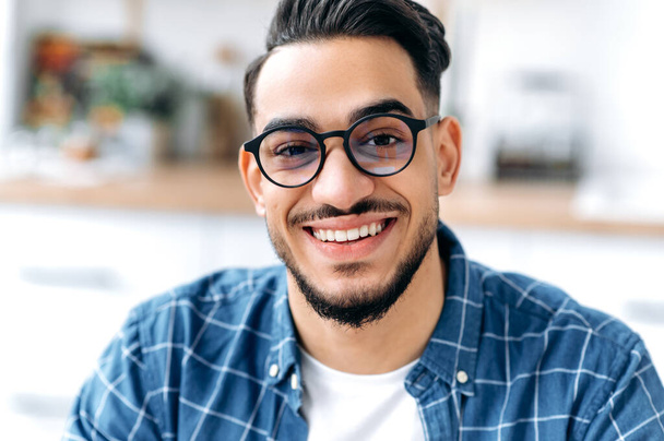 Nahaufnahme eines hübschen charismatischen arabischen oder indischen Typen mit Brille, Freiberufler oder Student, in lässiger Kleidung, mit perfekt weißen Zähnen, der mit einem zahmen, glücklichen Lächeln in die Kamera blickt - Foto, Bild
