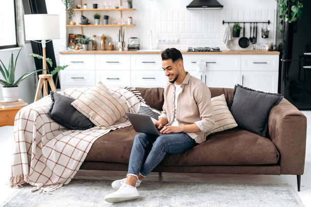 Positive handsome smart indian or arabian guy, freelancer or student, work or study online from home, using laptop, while sit on comfortable sofa in cozy living room, browsing internet, news, smiling - Fotoğraf, Görsel