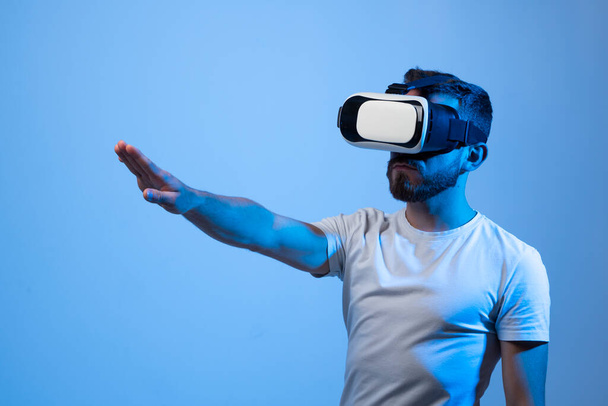 Hombre barbudo joven en casco de realidad virtual juega juego en línea en metaverso. Juego simula el comportamiento en el mundo ficticio, gadgets y adicción a la realidad virtual. Concepto de tecnología futura - Foto, imagen