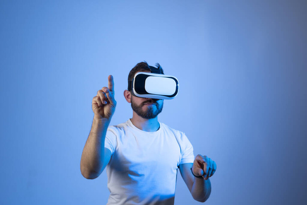 Young bearded man in virtual reality helmet plays online game in metaverse. Game simulates behavior in fictional world, gadgets and virtual reality addiction. Future technology concept - Fotoğraf, Görsel