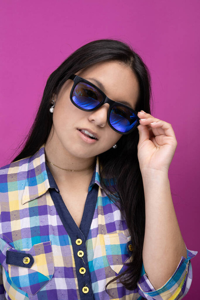 young latin girl with features, long black hair and a plaid shirt posing with her sunglasses, beauty and fashion in studio - Foto, Imagen