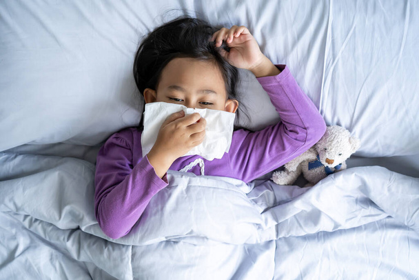 Top view Asian child girl sick with sneezing on the nose and cold cough on tissue paper because weak, Sick child with fever and illness in bed. - Фото, изображение
