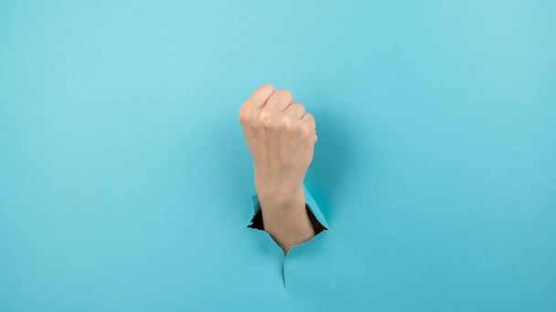 A womans hand sticking out of a hole from a blue background shows a fist - Foto, imagen