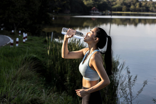 Koşudan sonra su içen Asyalı bir kadın, birçok insanın sabah ve akşam koşuya geldiği bir parkta koşuyor, koşmak popüler bir etkinliktir. Koşu ile sağlık konsepti. - Fotoğraf, Görsel