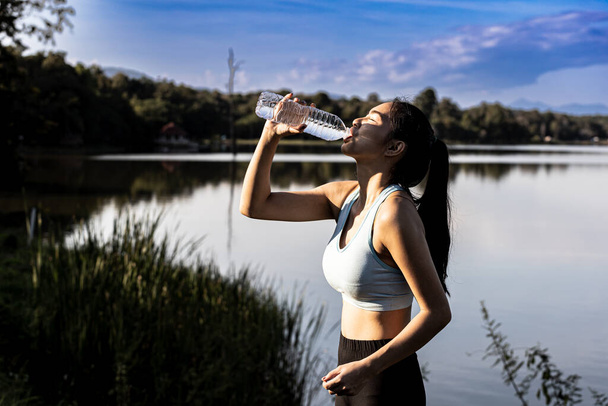 Femme asiatique boire de l'eau après le jogging, elle court dans un parc où beaucoup de gens viennent faire du jogging le matin et le soir beaucoup, la course est une activité populaire. Concept de soins de santé avec jogging. - Photo, image