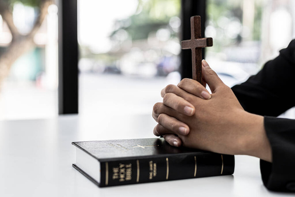 Bir insan İncil 'in kitabı üzerinde ellerini birleştirip bir haç tutarak İncil' e dua eder, okur ve incili okur. Hıristiyanlık ve İncil Çalışması kavramı. - Fotoğraf, Görsel