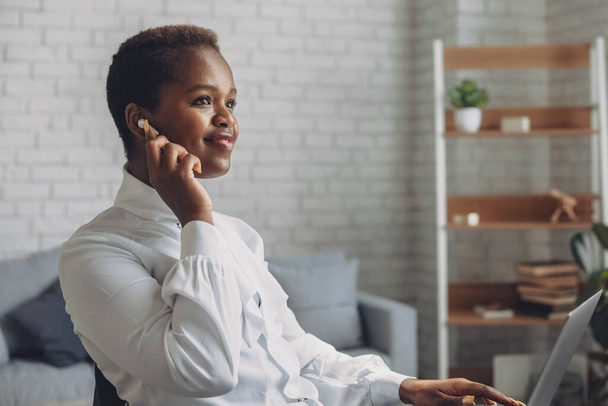 African-american business woman talking with earphone while checking some documents saved in her laptop. Digital marketing. Video call screen. Beautiful african - 写真・画像