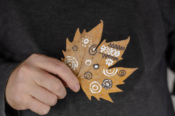 Een man houdt een gevallen blad met witte patronen vast. Witte abstracte patronen op een droog blad. Schilderen met verf op gevallen bladeren. Hedendaagse kunst. Selectieve focus. - Foto, afbeelding