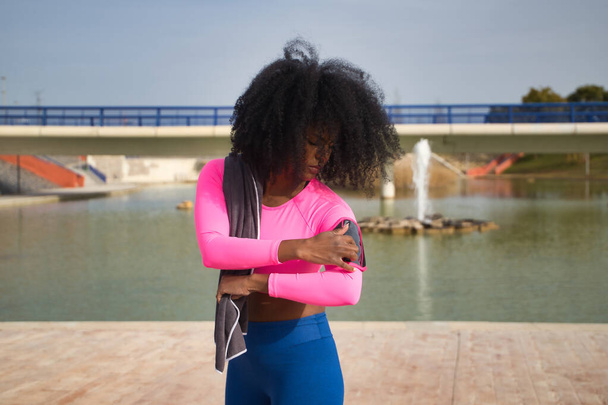 Mulher afro-americana com cabelo afro e roupas esportivas, vestindo camiseta rosa fluorescente e leggings, abrindo um aplicativo em execução em seu telefone celular. Conceito de aptidão, esporte, rua, corrida. - Foto, Imagem