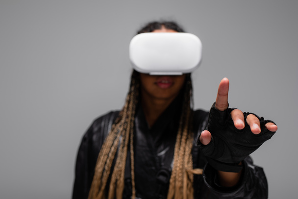Mulher afro-americana desfocada em fone de ouvido de realidade virtual apontando com o dedo isolado em cinza  - Foto, Imagem