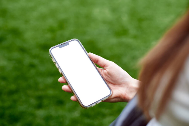 Het meisje houdt de telefoon vast met een leeg scherm, zicht van achteren. Adverteer uw app. Mockup afbeelding van de hand te houden witte mobiele telefoon met blanco wit. - Foto, afbeelding