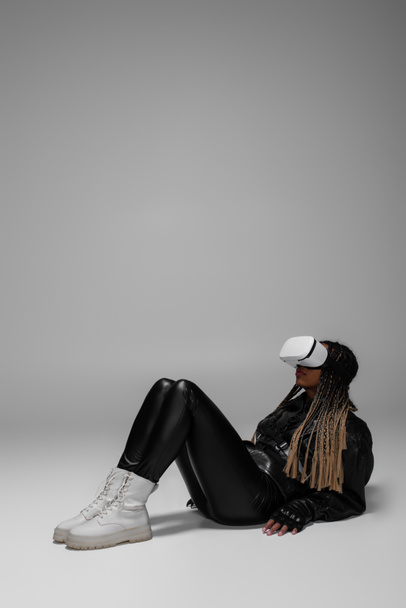 African american woman in leather jacket and boots using vr headset on grey background  - Φωτογραφία, εικόνα