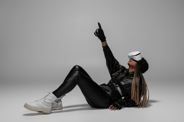 African american woman in vr headset and gloves pointing with finger while sitting on grey background  - Φωτογραφία, εικόνα