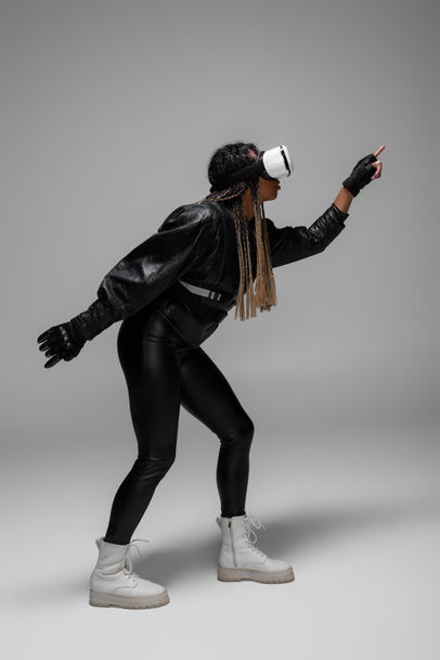 Vista lateral de la mujer afroamericana en auriculares de realidad virtual y juegos de chaqueta de cuero sobre fondo gris  - Foto, Imagen