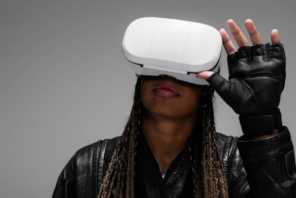Αφροαμερικανή γυναίκα με δερμάτινο γάντι χρησιμοποιώντας ακουστικά VR απομονωμένα σε γκρι  - Φωτογραφία, εικόνα