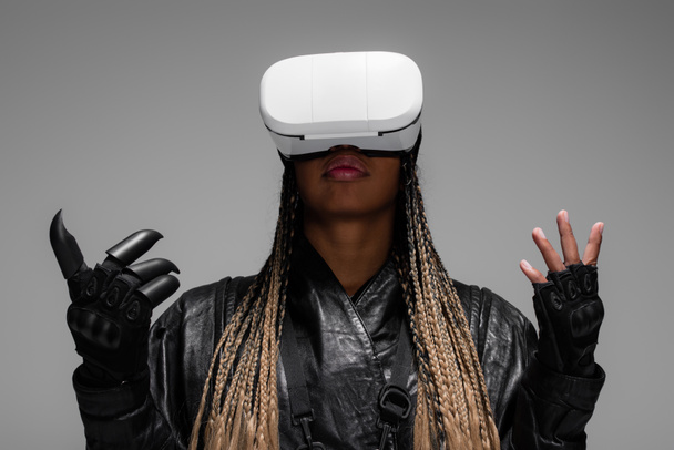 Afro-Amerikaanse vrouw in handschoenen en leren jas met behulp van virtual reality headset geïsoleerd op grijs  - Foto, afbeelding