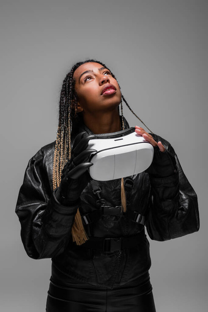 Trendy african american woman holding vr headset and looking up isolated on grey  - Zdjęcie, obraz