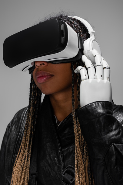 Африканская женщина с роботизированной рукой и VR-гарнитурой слушает музыку в VR-гарнитуре, изолированной на сером фоне  - Фото, изображение