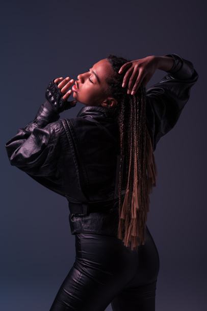 African american woman in glove and leather jacket touching pigtails isolated on dark grey  - Zdjęcie, obraz