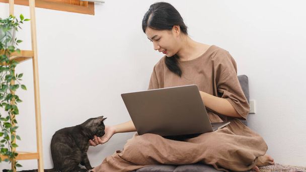 Kisállat szerető koncepció, fiatal ázsiai nő használ laptop és játszik simogató macskával a nappaliban. - Fotó, kép