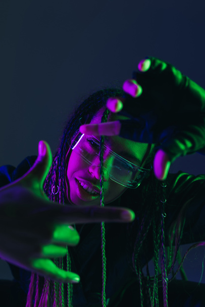 Smiling african american woman in smart glasses gesturing isolated on dark blue  - Foto, imagen