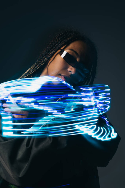 Retrato de mujer afroamericana con estilo en gafas inteligentes posando cerca de luces de neón azules sobre fondo gris  - Foto, Imagen