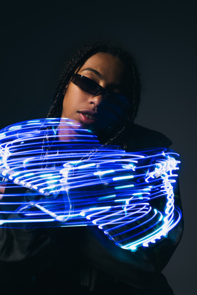 Trendy african american woman in smart glasses posing near abstract blue lights on grey background  - Photo, Image