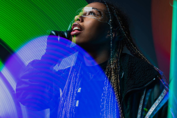 Африканская американка в умных очках и кибер-перчатках смотрит в сторону абстрактных огней на сером фоне  - Фото, изображение
