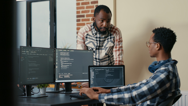 Software developer sitting at desk with multiple screens and laptop running code talking with colleague about artificial intelligence algorithm. Software developers doing teamwork in it office. - Photo, image