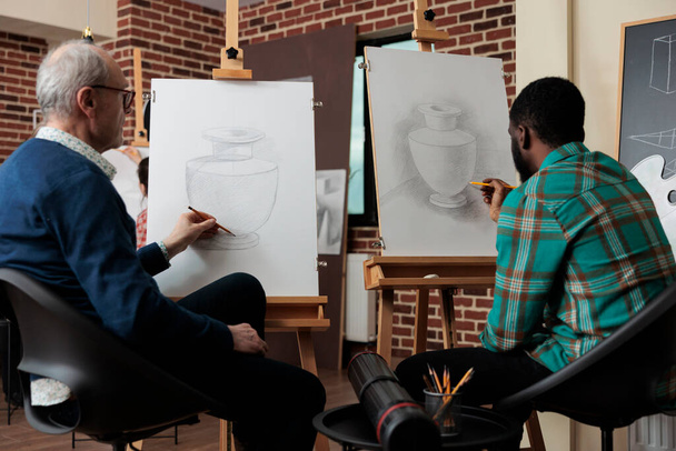 Yaratıcılık stüdyosundaki resim dersinde çizim teknikleri üzerinde çalışan grafiksel kalem kullanarak tablo üzerinde vazo modeli çizen çok ırklı öğrenciler. Çeşitli takımlar yeni sanatsal yetenekler geliştiriyor. - Fotoğraf, Görsel