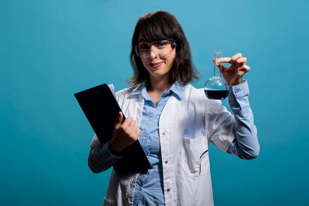 Microbiology expert on blue background holding glass beaker filled with chemical compound. Biochemistry research specialist holding volumetric glass flask filled with unknown liquid substance sample. - Photo, Image