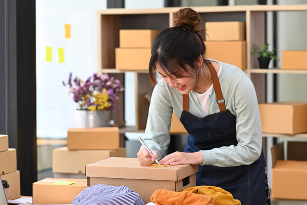Online-Verkäuferin schreibt Adresse auf Karton, während sie zu Hause im Büro arbeitet. Konzept des Online-Verkaufs, E-Commerce. - Foto, Bild