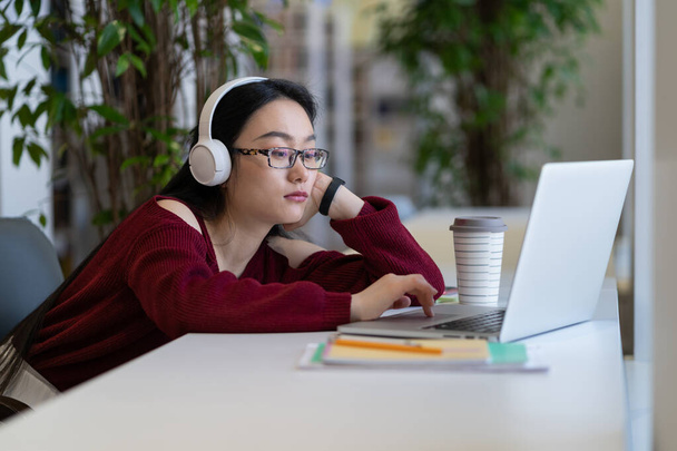 Устала скучать азиатская студентка в наушниках, пользующаяся интернетом на ноутбуке во время учебы в современной университетской библиотеке, избирательная направленность. Промедление в онлайн-обучении, дистанционном обучении - Фото, изображение