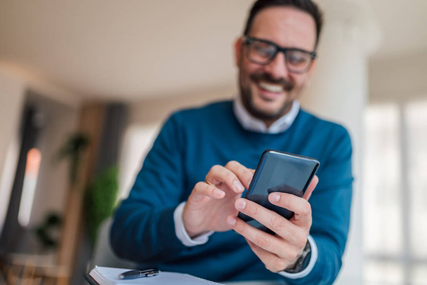 Close-up of young businessman text messaging on mobile phone. Smiling male executive is wearing smart casuals at office. Focus is on smart phone and hands.. - Zdjęcie, obraz