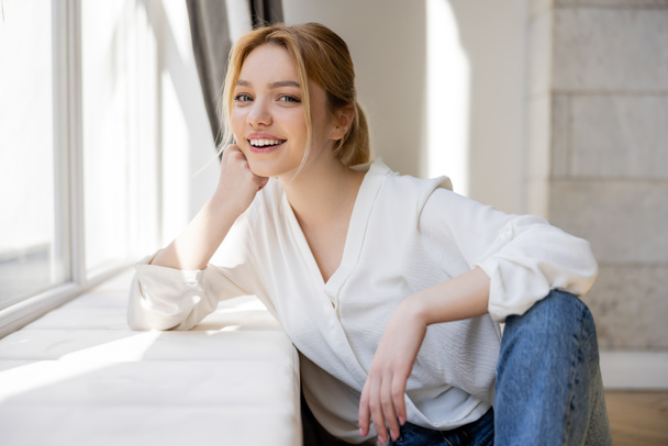 Portrait of smiling blonde woman looking at camera near windowsill at home  - Photo, Image