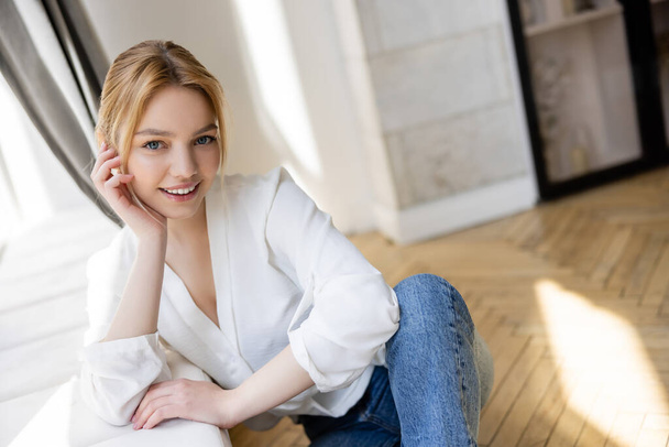 Pretty blonde woman in blouse looking at camera near windowsill at home  - Foto, Imagem