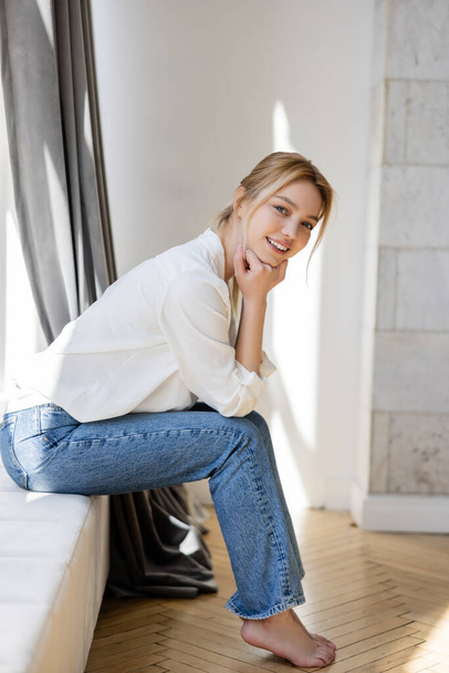 Mujer descalza positiva en jeans y blusa mirando la cámara en el alféizar de la ventana en casa  - Foto, imagen
