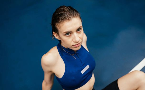 Portrait of a beautiful sports woman in sportswear lying on a wet sports field during a workout in the rain and looking at the camera on a blue background - Zdjęcie, obraz
