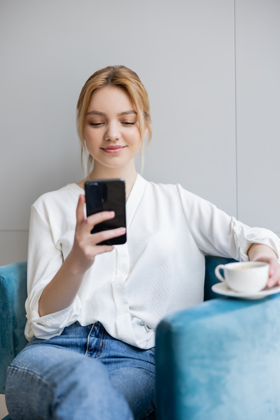 Positive blonde woman using blurred smartphone and holding cup on armchair at home  - Foto, Bild