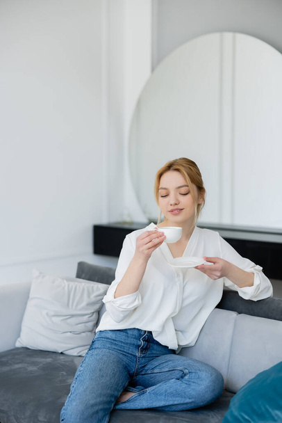Blonde woman in jeans and blouse holding cup of coffee in living room  - Valokuva, kuva