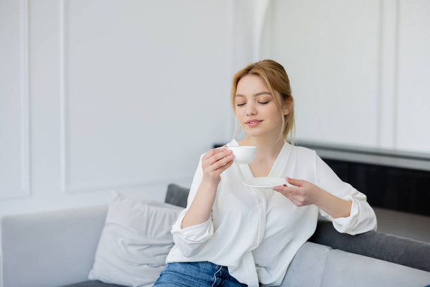Pretty blonde woman holding cup and saucer on couch at home  - Foto, Bild