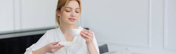 Young woman in blouse holding coffee cup and saucer in living room, banner  - Foto, imagen