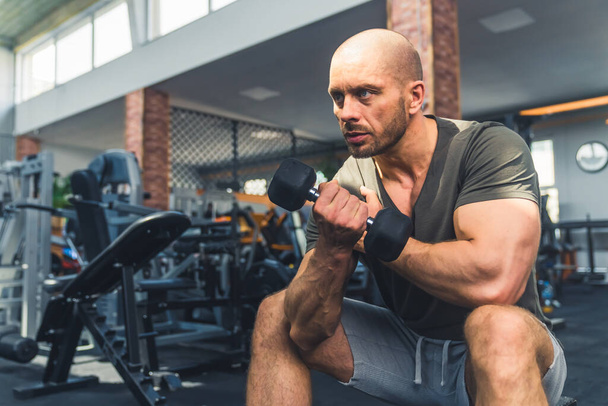 Hombre musculoso serio concentrado en sus 30 años sentado en un banco de gimnasio mirando hacia adelante, sosteniendo una pequeña mancuerna en su mano. Foto de alta calidad - Foto, Imagen