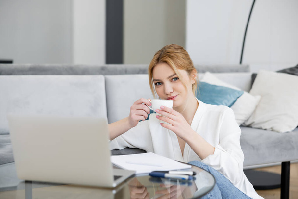 Blonde freelancer holding cup near blurred laptop and notebook on coffee table at home  - Photo, Image