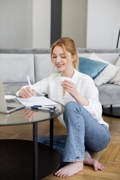 Hübsche blonde Freiberuflerin hält Tasse in der Hand und schreibt auf Notizbuch neben Laptop auf Couchtisch im Wohnzimmer  - Foto, Bild