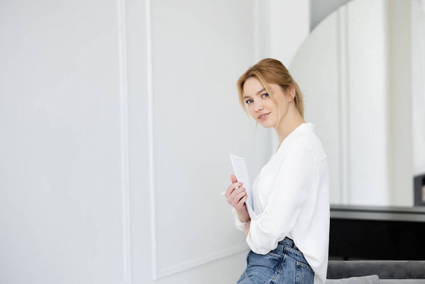 Happy young woman holding notebook and looking at camera in living room  - Foto, afbeelding