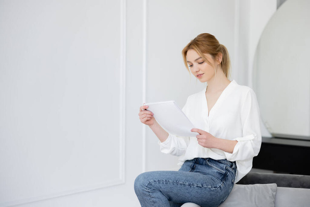 Mujer joven en blusa sosteniendo cuaderno en el sofá en casa  - Foto, Imagen