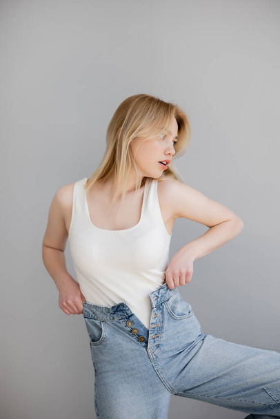 Blonde woman in bodysuit and jeans looking away isolated on grey  - Foto, imagen