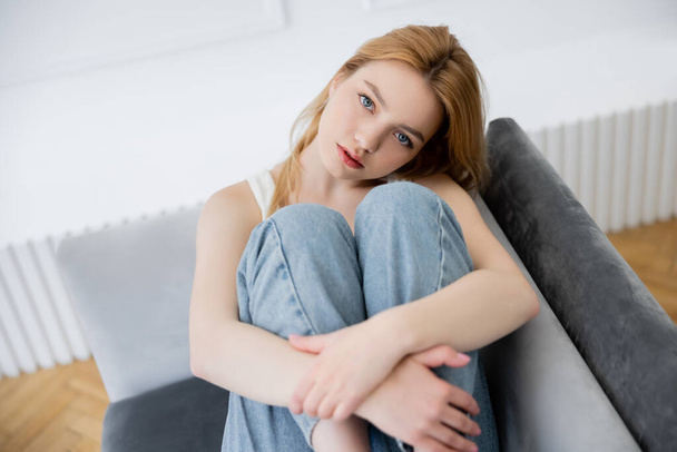 Portret van jonge vrouw in wazig jeans kijkend naar camera terwijl ze op de bank zit  - Foto, afbeelding
